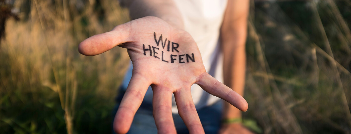 Not sehen und handeln Eine Person, die ein weißes T-Shirt trägt und in einer Sommerwiese steht, streckt eine Hand mit Schriftzug "Wir helfen" in die Kamera.
